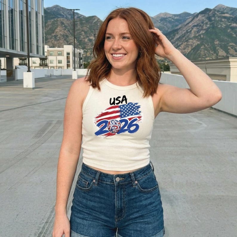 May include: A cream-colored ribbed tank top featuring a graphic print with the text USA, a stylized American flag design, and the year 2026 in bold blue font, paired with dark blue denim shorts with a visible button and zipper fly closure.