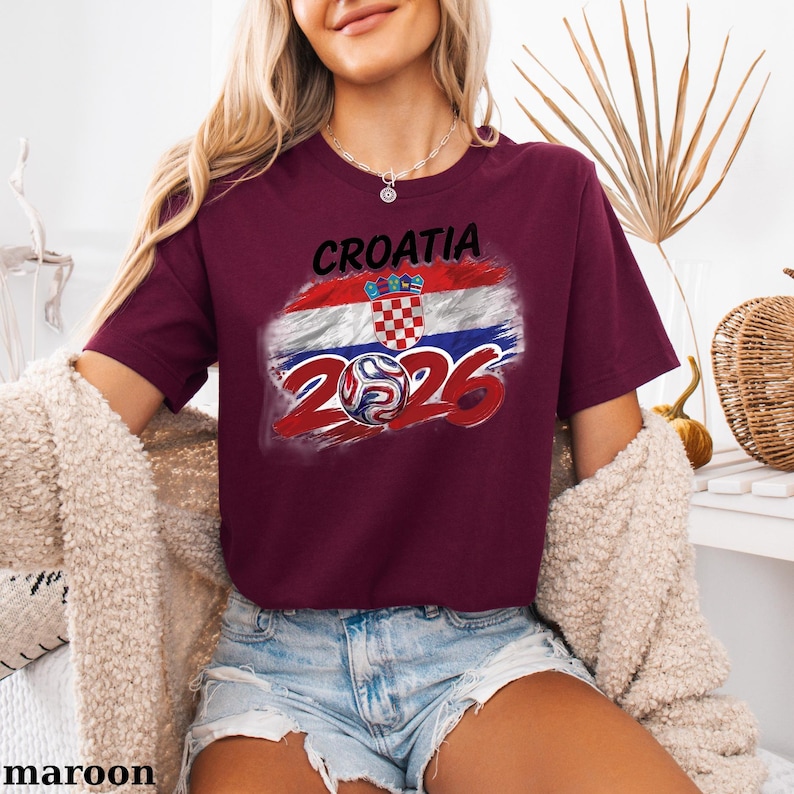 May include: Maroon t-shirt with the word "CROATIA" above a graphic of the Croatian flag and a football with the numbers "2026". The shirt has short sleeves and a crew neck.