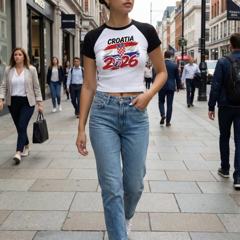 May include: A white and black raglan crop top featuring a graphic print of the Croatian flag with the text Croatia 2026 and a football icon. The shirt has short black sleeves and a white body, paired with light blue denim jeans.