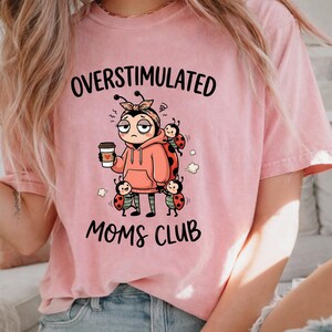 May include: Pink t-shirt with the words "OVERSTIMULATED MOMS CLUB" in black. The graphic features a cartoon ladybug character wearing a pink hoodie and holding a coffee cup, surrounded by other ladybugs.