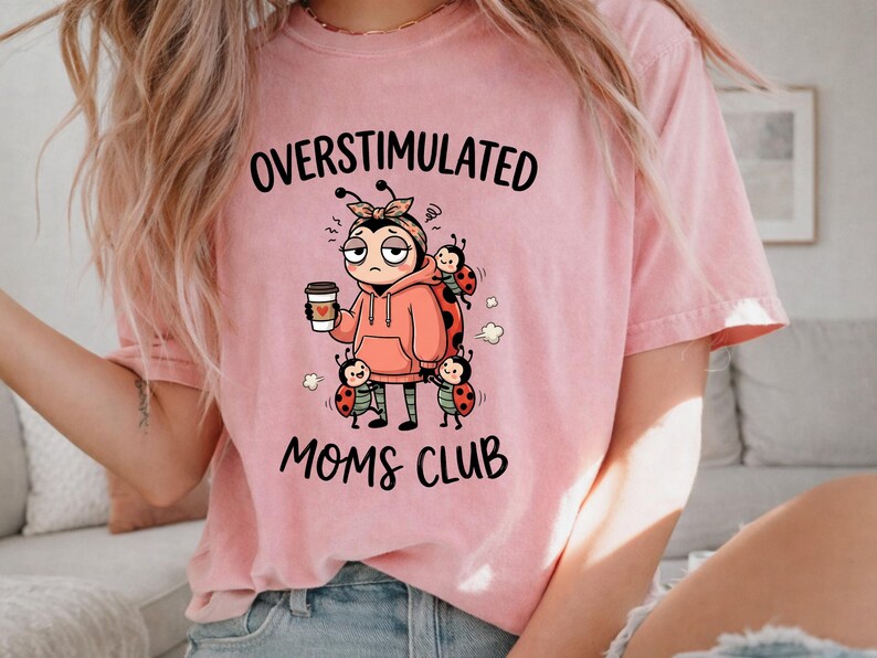May include: Pink t-shirt with the words "OVERSTIMULATED MOMS CLUB" in black. The graphic features a cartoon ladybug character wearing a pink hoodie and holding a coffee cup, surrounded by other ladybugs.