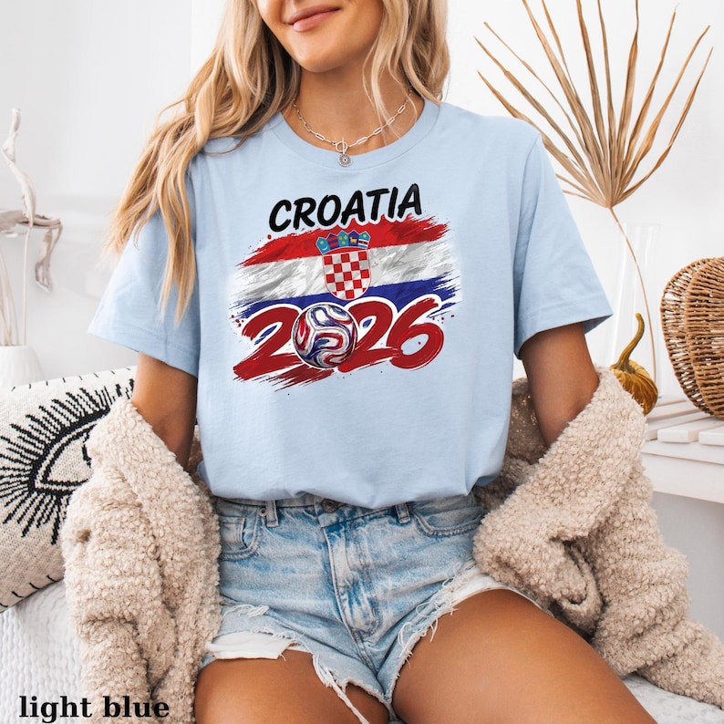 May include: Pale blue t-shirt with the word "CROATIA" above a design of the Croatian flag and a soccer ball with the number "2026" in red. The shirt is made of a soft material.