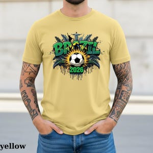 May include: Yellow t-shirt with a Brazil-themed graphic. The design features the word "BRAZIL" in green, a soccer ball, and the year "2026". The graphic includes palm leaves and a depiction of Christ the Redeemer statue.