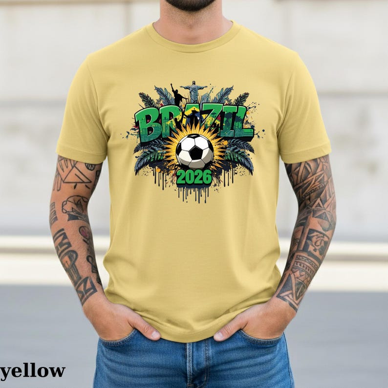 May include: Yellow t-shirt with a Brazil-themed graphic. The design features the word "BRAZIL" in green, a soccer ball, and the year "2026". The graphic includes palm leaves and a depiction of Christ the Redeemer statue.