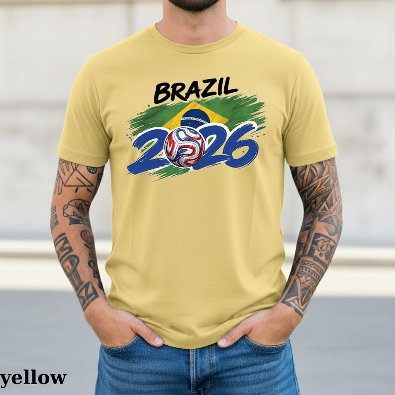Pu&ograve; includere: Una maglietta gialla con la scritta "BRAZIL" in nero sopra una grafica della bandiera brasiliana e il numero "2026" in blu. Una grafica di pallone da calcio &egrave; al centro del "2026".