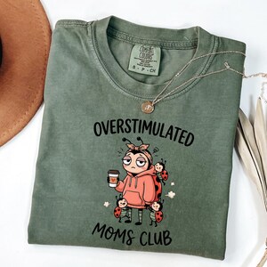 May include: Olive green t-shirt with the words "OVERSTIMULATED MOMS CLUB" and a cartoon ladybug graphic. The ladybug is wearing a pink hoodie and holding a coffee cup. A silver necklace is draped across the shirt.