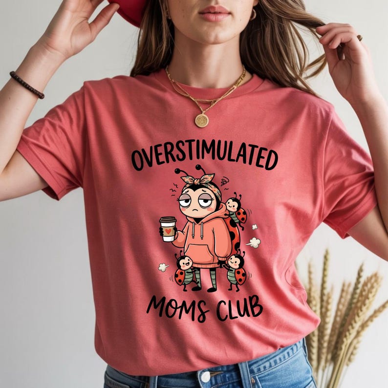 May include: Coral-colored t-shirt with the words "OVERSTIMULATED MOMS CLUB" in black. The graphic features a cartoon ladybug character holding a coffee cup, surrounded by other ladybugs. The ladybug character is wearing a hoodie and has a tired expression.