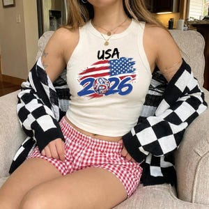 May include: A cream-colored racerback tank top featuring a graphic print with the text USA, a stylized American flag, and the number 2026 with a circular star-patterned emblem. The tank top is worn with red and white gingham shorts and a black and white checkered cardigan.