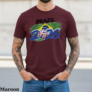 Pu&ograve; includere: T-shirt bordeaux con un design Brasile 2026. Il design presenta la parola "BRAZIL" in nero, uno sfondo verde e giallo, il numero "2026" in blu e un pallone da calcio.