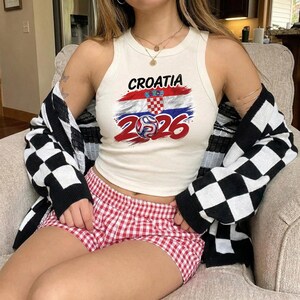 May include: A cream-coloured sleeveless tank top featuring a graphic of the Croatian flag, a football, and the text Croatia 2026. The top is worn with red and white gingham shorts and a black and white chequered long-sleeve cardigan sweater.