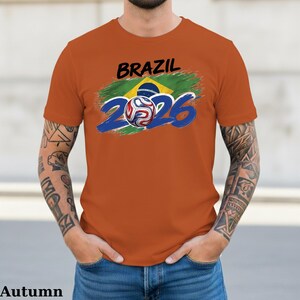 Pu&ograve; includere: T-shirt arancione con la scritta "BRAZIL" in nero sopra una grafica della bandiera brasiliana e il numero "2026" in blu. Un pallone da calcio &egrave; al centro del design "2026".