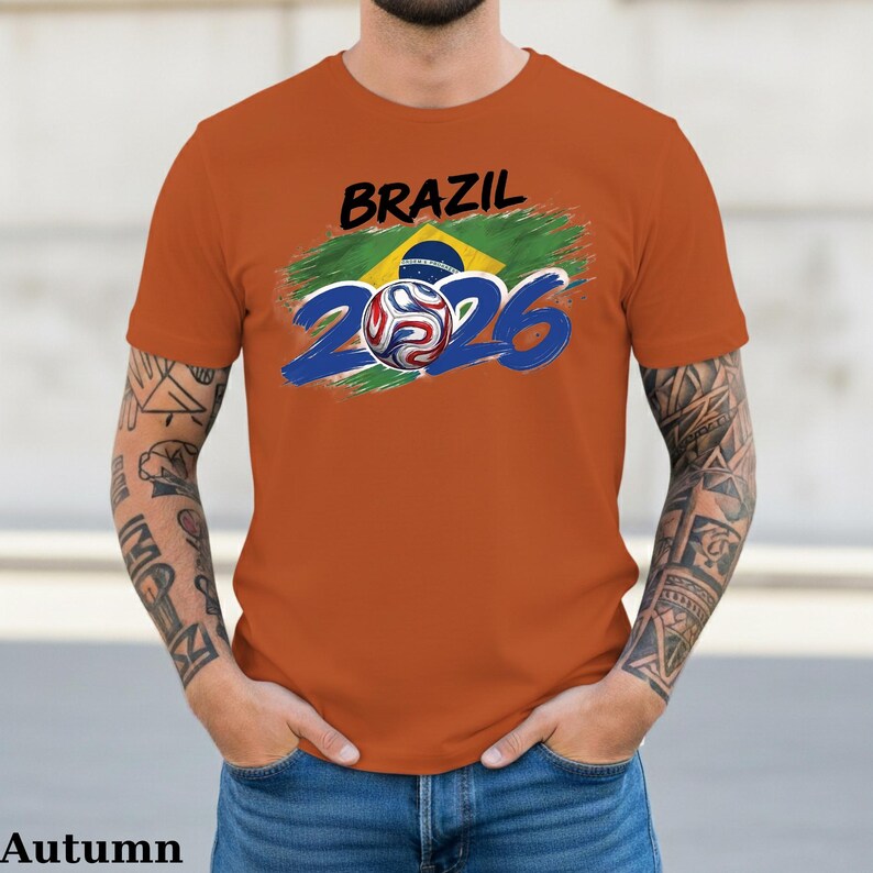 Pu&ograve; includere: T-shirt arancione con la scritta "BRAZIL" in nero sopra una grafica della bandiera brasiliana e il numero "2026" in blu. Un pallone da calcio &egrave; al centro del design "2026".