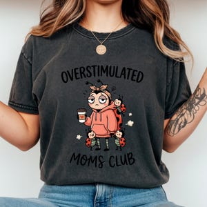 May include: Dark gray t-shirt with the words "OVERSTIMULATED MOMS CLUB" and a cartoon ladybug design. The ladybug character is wearing a pink hoodie and holding a coffee cup. Several other ladybugs are also featured.