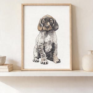 Könnte beinhalten: Aquarelldruck eines Deutsch Kurzhaar Welpen, gerahmt in hellem Holz. Der Welpe ist sitzend dargestellt, mit braunen und grauen Markierungen und Schlappohren. Das Kunstwerk wird auf einem weißen Regal mit dekorativen Gegenständen ausgestellt.