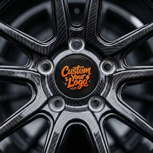 Könnte beinhalten: Nahaufnahme eines schwarz-grauen Autorads mit einem Kohlefaserdesign. Die Radkappe ist schwarz mit orangefarbenem Text, der "Custom Your Logo" lautet. Das Rad hat ein sternförmiges Speichendesign.