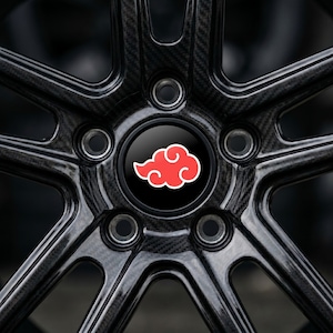 May include: Close-up of a black car wheel with a carbon fiber texture. The wheel's center cap features a black background with a red cloud design outlined in white. The wheel has a five-spoke design.