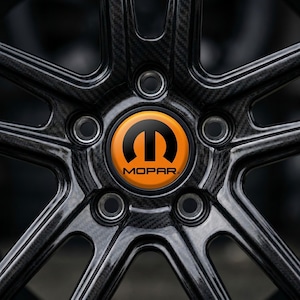 May include: Close-up of a black car wheel with a carbon fiber weave pattern. The center cap is orange with the black Mopar logo and text. The wheel has a multi-spoke design.