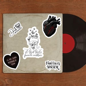 May include: A collection of stickers on a vintage record sleeve. The stickers feature gothic and romantic themes, including a black heart with red veins, a skull crown, and text that reads "Dark Romance" and "A Good Girl's favorite necklace."