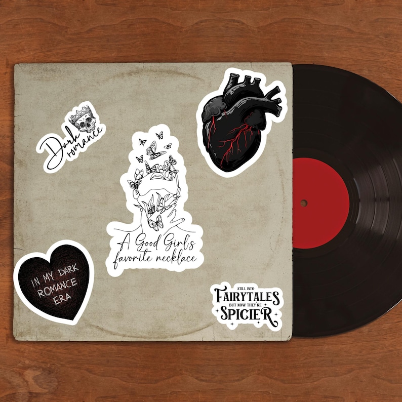May include: A collection of stickers on a vintage record sleeve. The stickers feature gothic and romantic themes, including a black heart with red veins, a skull crown, and text that reads "Dark Romance" and "A Good Girl's favorite necklace."
