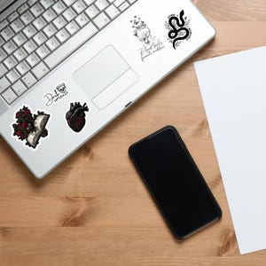 May include: A collection of stickers with gothic and romantic themes. Designs include a snake, a human heart, roses, and the text "Dark Romance" and "A Good Girl's favorite necklace". The stickers are on a laptop and a wooden surface.