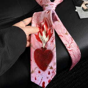 May include: A light pink necktie featuring a central graphic of a dark red heart pierced by two arrows with flames rising from the top. The fabric is decorated with a repeating pattern of small gold stars and red coral-like branch motifs.