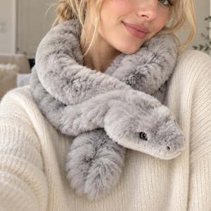 May include: A plush, light grey faux fur scarf shaped like a snake. The scarf features a detailed head with dark eyes and a textured, soft surface that wraps around the neck, mimicking the appearance of a coiled serpent in a neutral tone.