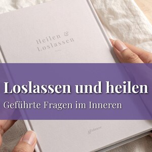 May include: A light gray book with the German text "Heilen & Loslassen" and "Loslassen und heilen" in white. The book is held by two hands, resting on a white cloth. The book's title translates to "Letting go and healing".