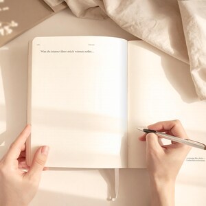 May include: An open journal with blank pages, held by two hands. A pen is poised over the right page, ready to write. The text "Was du immer &uuml;ber mich wissen solltest..." is visible. The scene is bathed in soft, natural light.