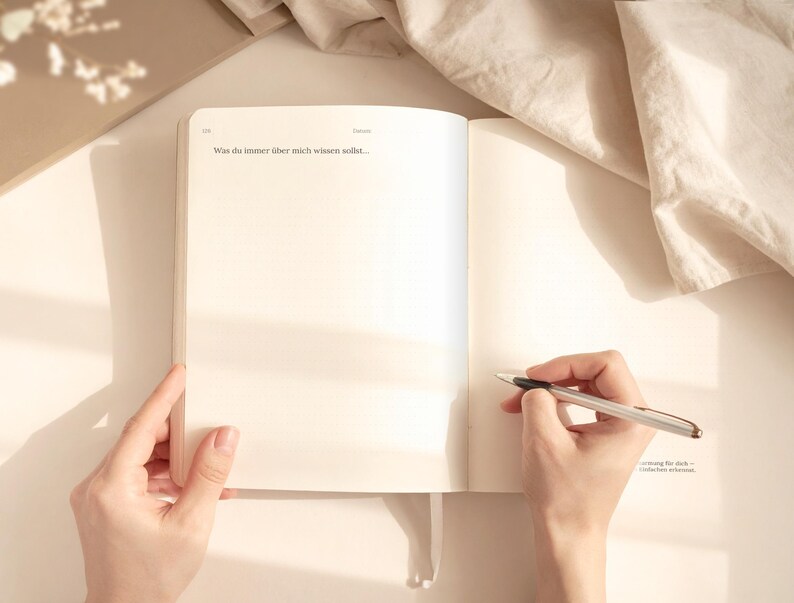 May include: An open journal with blank pages, held by two hands. A pen is poised over the right page, ready to write. The text "Was du immer &uuml;ber mich wissen solltest..." is visible. The scene is bathed in soft, natural light.