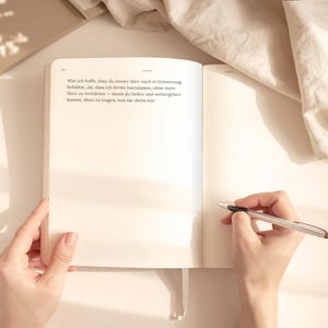 May include: An open notebook with text on the left page and blank on the right. A silver pen is held over the blank page. The notebook is on a light-colored surface, with a soft cloth in the background.
