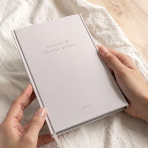 May include: A light gray journal with the words "Guérir & lâcher prise" printed on the cover. The journal has a ribbon bookmark and is held by two hands.