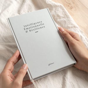 May include: A light gray journal with the words "Intelligence Émotionnelle & Résilience" printed on the cover. The journal has a matching gray bookmark and is held by two hands.