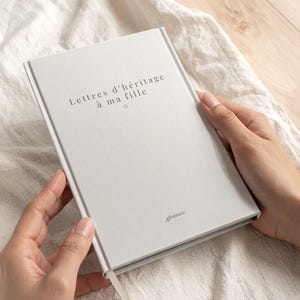 May include: A light gray journal with the text "Lettres d'héritage à ma fille" is held open. The journal has a matching ribbon bookmark and the logo "AEmbraces" on the bottom right corner. The journal is on a white, textured surface.