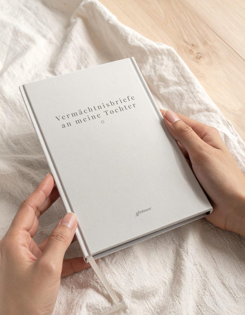May include: A light gray journal with the German text "Verm&auml;chtnisbriefe an meine Tochter" is held open. The journal has a ribbon bookmark and the logo "A Embraces" at the bottom right.