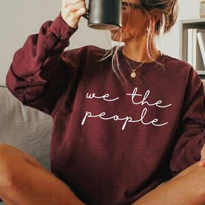 May include: A maroon sweatshirt with the text "we the people" in white script. The person is holding a black mug. The person is wearing a gold necklace and brown slippers.