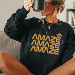 May include: Black sweatshirt with the word "AMAZE" in yellow and orange lettering. The word is repeated three times with a planet graphic. The person is holding a black mug.