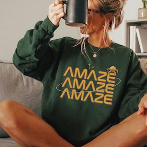 May include: A dark green sweatshirt with the word "AMAZE" in yellow, repeated three times with a planet-like design. The person is holding a black mug. The person is wearing brown slippers with white fur.