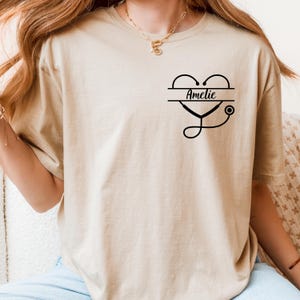 May include: A beige t-shirt featuring a black heart-shaped design with the name "Amelie" and a stethoscope graphic. The shirt is made of a soft material and has a classic crew neck.
