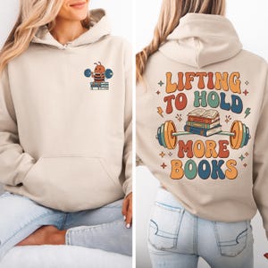 May include: Beige hoodie with a front pocket and a graphic of a bee lifting weights over books. The back of the hoodie features the text "Lifting to Hold More Books" in a retro font with a barbell and books graphic.