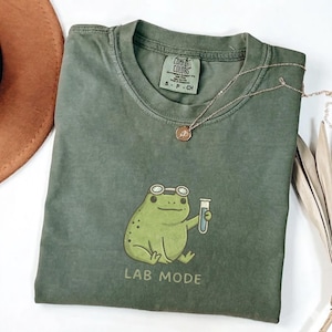 Puede incluir: Camiseta verde oliva con un dibujo de una rana con gafas y un tubo de ensayo. El texto "LAB MODE" está impreso debajo de la rana. También se ven un collar dorado y un sombrero marrón.