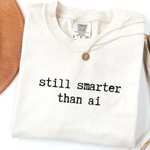May include: Cream-colored t-shirt with the text "still smarter than ai" in black. A gold necklace with a circular pendant rests on the shirt. A brown hat is visible in the upper left corner.
