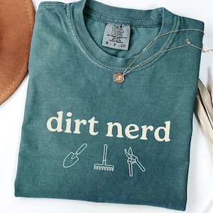 Puede incluir: Camiseta Comfort Colors de color verde azulado con las palabras "dirt nerd" e ilustraciones de herramientas de jardinería. Un collar dorado con un colgante descansa sobre la camiseta. Un sombrero marrón es parcialmente visible.