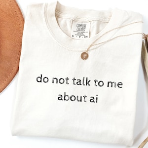 Peut inclure: T-shirt crème avec le texte "do not talk to me about ai" en noir. Un collier doré avec un pendentif circulaire repose sur le t-shirt. Un chapeau marron est partiellement visible dans le coin supérieur gauche.