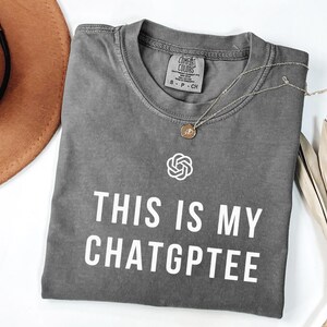 Op de afbeelding: Een grijze t-shirt met de tekst "THIS IS MY CHATGPTEE" in witte blokletters. De t-shirt heeft een ronde hals en korte mouwen. Een zilveren ketting met een hanger ligt op de t-shirt. De t-shirt is op een witte ondergrond gevouwen.