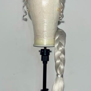 Könnte beinhalten: Silberblondes Perücke mit lockeren Locken und einem langen, geflochtenen Pferdeschwanz. Der Zopf ist mit kleinen, funkelnden Verzierungen versehen und mit einem dekorativen Verschluss befestigt. Die Perücke wird auf einem Schaufensterkopf präsentiert.