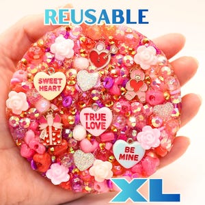 Puede incluir: Un posavasos circular, reutilizable y XL de resina, incrustado con cuentas, lentejuelas y dijes rosas, rojos e iridiscentes. Los dijes incluyen un osito de peluche, un regalo y piezas en forma de corazón con el texto SWEET HEART, TRUE LOVE, BE MINE.