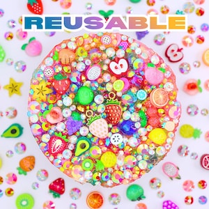 May include: A circular, bedazzled object with fruit-shaped embellishments, including strawberries, avocados, and lemons. The word "REUSABLE" is displayed across the top. The object is surrounded by scattered, colourful rhinestones.