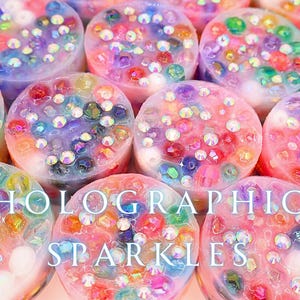 Op de afbeelding: Close-up van verschillende ronde, kleurrijke objecten met holografische sparkles. De objecten zijn een mix van roze, paars, blauw en groen, met ingebedde iriserende edelstenen. De tekst "HOLOGRAPHIC SPARKLES" staat over de afbeelding.