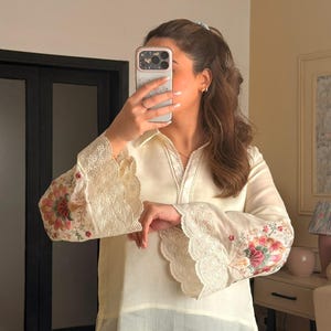 May include: A cream-coloured long-sleeve shirt featuring a pointed collar and a V-neckline. The wide cuffs and the bottom hem are finished with intricate white eyelet embroidery. The sleeves display colourful floral embroidery in pink and green.