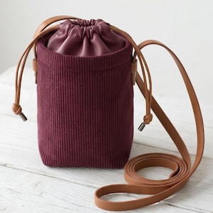 May include: A cylindrical bucket bag featuring a deep burgundy corduroy exterior and a matching burgundy drawstring closure. The bag includes a long, thin tan leather shoulder strap attached to the sides, providing a functional carry option.